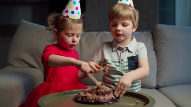 Dos niños en sombreros de fiesta comiendo pastel de chocolate de cumpleaños con cucharas y manos de madera. Hermano y hermana disfrutando comiendo dulces en casa . — Vídeos de Stock