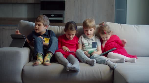 Grupo de cuatro niños de edad mixta que utilizan gadgets sentados juntos en el sofá en casa. Hermanos jugando y viendo en línea en teléfonos móviles y tabletas. Dos chicos y dos chicas con dispositivos . — Vídeos de Stock