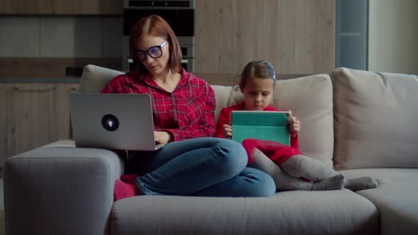 Trabajando desde casa mujer de 30 años usando el ordenador portátil con su hija usando la tableta de la computadora sentada en el sofá en casa. Familia usando artilugios en casa. Trabajo y estudio en línea . — Vídeo de stock