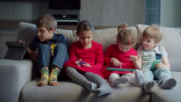 Een groep van vier kinderen van gemengde leeftijd die gadgets gebruiken die thuis op de bank zitten. Broers en zussen spelen en kijken online op mobiele telefoons en tablet computers. Twee jongens en twee meisjes met apparaten. — Stockvideo