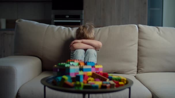 Niño rubio preescolar trastornado sentado en el sofá en casa. Niño insatisfecho con constructor de color bloques de juguete en la mesa . — Vídeos de Stock