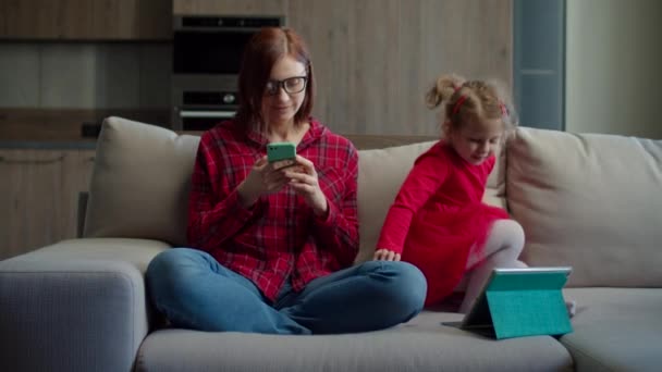30 'lu yaşlarda, gözlüklü bir anne, küçük bir kızla cep telefonu kullanıyor. Evdeki kanepede oturan tablet bilgisayar kullanıyor. Aile aletleri birlikte kullanıyor.. — Stok video