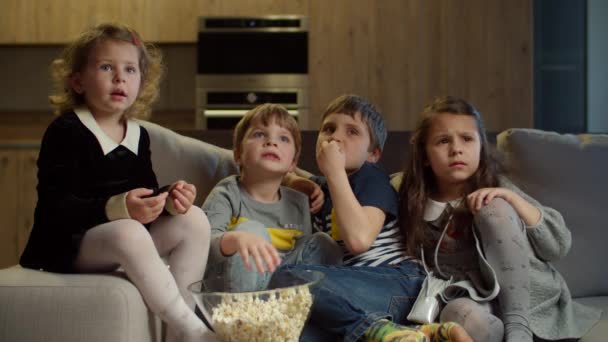 Grupo de cuatro niños viendo la televisión, comiendo palomitas de maíz sentados en el sofá en casa. Dos chicas y dos chicos viendo películas emocionantes en casa. Hermanos en el sofá . — Vídeo de stock