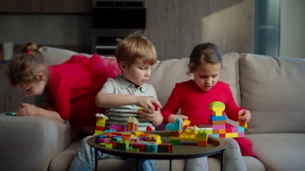 Dvě děti si hrají s barevnými konstruktérskými bloky a jedna holčička používá mobilní telefony, které sedí doma na gauči. Sourozenci hrají spolu. — Stock video