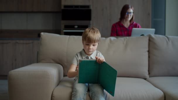 Niño preescolar usando tableta de computadora para la educación en línea sentado en el sofá con la madre trabajando desde casa en el ordenador portátil detrás. Familia con gadgets de trabajo y estudio en línea en casa . — Vídeo de stock