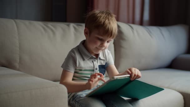 Νηπιαγωγείο χρησιμοποιώντας τον υπολογιστή tablet για online εκπαίδευση κάθεται στον καναπέ στο σπίτι. Παιδί που χρησιμοποιεί την εφαρμογή για να μελετήσει online. Παιδί με gadget. — Αρχείο Βίντεο