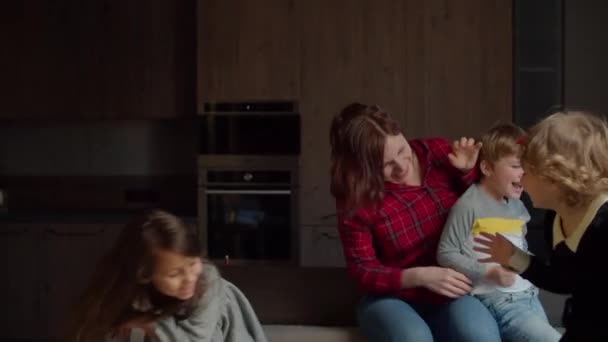 Trójka dzieci skacze i tańczy z matką na kanapie w ciemnym pokoju. Chłopiec i dziewczyny dobrze się bawią z mamą na sofie. — Wideo stockowe