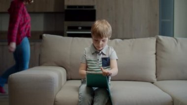 Anaokulu çocuğu online eğitim için tablet bilgisayar kullanıyor. Annesiyle birlikte kanepeye oturuyor ve bilgisayarını açıyor. Cihazları olan bir aile evde internetten çalışıyor ve okuyor.. 