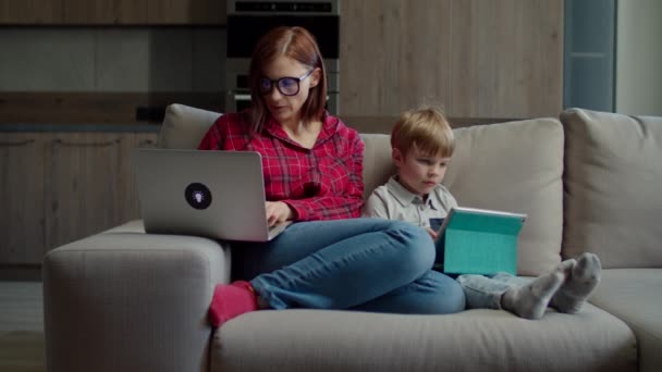 Trabalhando a partir de casa jovem 30 mãe com laptop e menino pré-escolar com computador tablet sentado no sofá em casa. Família usando gadgets para educação on-line e trabalho — Vídeo de Stock