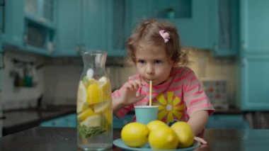 Pembe tişörtlü, kağıt kamışlı ev yapımı limonata içen, mavi mutfakta duran sevimli anaokulu kızı. Masada sarı limon meyveleri var.. 