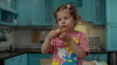 Pembe tişörtlü, ev yapımı pizza yiyen sevimli küçük bir kız. Evdeki mavi mutfakta duruyor. Çocuk elleriyle pizza yemekten hoşlanıyor.. 