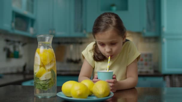 Menina da escola em vestido amarelo beber copo de limonada caseira com palha de papel, de pé na cozinha azul em casa. Frutas de limão amarelas na mesa . — Vídeo de Stock