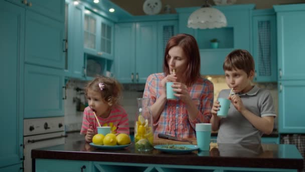 Madre joven con dos hijos bebiendo limonada casera de pie en la cocina azul en casa. Mamá con hijo e hija bebiendo vaso de bebida de limón con paja de papel . — Vídeos de Stock