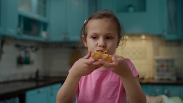Niña preescolar en camiseta rosa comiendo pizza casera de pie en la cocina azul en casa. Niño disfrutar comiendo pizza con las manos . — Vídeos de Stock
