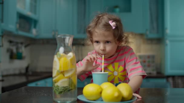 Roztomilá školačka v růžovém tričku popíjející sklenici domácí limonády s papírovou slámou, stojící doma na modré kuchyni. Žluté citrónové plody na stole. — Stock video