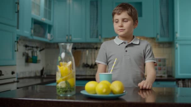 Niño de la escuela en camiseta gris beber vaso de limonada casera con paja de papel, de pie en la cocina azul en casa. Frutas amarillas de limón sobre la mesa . — Vídeos de Stock
