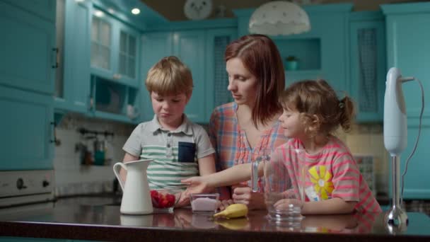 Feliz familia de madre y dos hijos cocinando batido de fresas, plátano, leche vegana y yogur con licuadora en casa cocina azul. Niño y niña vertiendo frutas en un tazón . — Vídeos de Stock