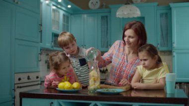 Üç çocuklu bir aile ev yapımı limonata, nane ve soda yapıyor. Genç anne, mavi mutfakta çocuklarla yemek pişiriyor.