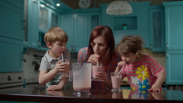 Gelukkige familie van moeder en twee kinderen drinken bessen smoothie met papieren stro thuis blauwe keuken. Kinderen genieten van zelfgemaakte milkshake. — Stockvideo