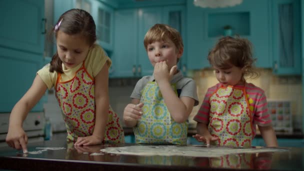 Tre bambini in grembiuli che giocano con la pasta sul tavolo della cucina a casa. I bambini assaggiano la pasta cruda leccando le dita. Fratelli che si divertono insieme in cucina . — Video Stock