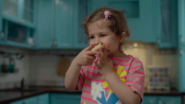 Linda niña en camiseta rosa comiendo pizza casera de pie en la cocina azul en casa. Niño disfrutar comiendo pizza con las manos . — Vídeo de stock