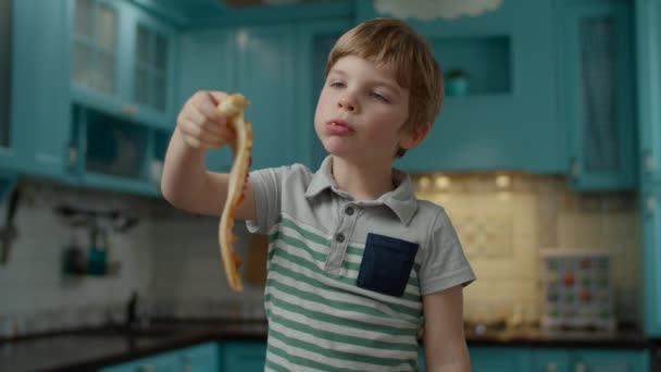自宅の青いキッチンに立つ自家製ピザを食べるTシャツの就学前の男の子。子供たちは手でピザを食べる. — ストック動画
