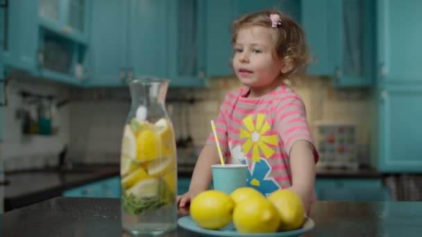 ピンクのTシャツに身を包んだ可愛い就学前の女の子は、自家製レモネードのグラスを紙の藁で飲み、自宅の青いキッチンに立っています。テーブルの上に黄色いレモンの果物. — ストック動画