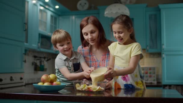 Feliz familia de madre y dos hijos cocinando tarta de manzana junto con huevos, azúcar, harina y manzanas. Niño y niña vertiendo masa en un tazón con manzanas . — Vídeos de Stock