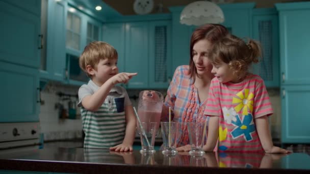 Familia feliz de la madre y dos niños bebiendo batido de bayas en casa cocina azul.Mujer vertiendo cóctel de fresa saludable a los niños . — Vídeos de Stock