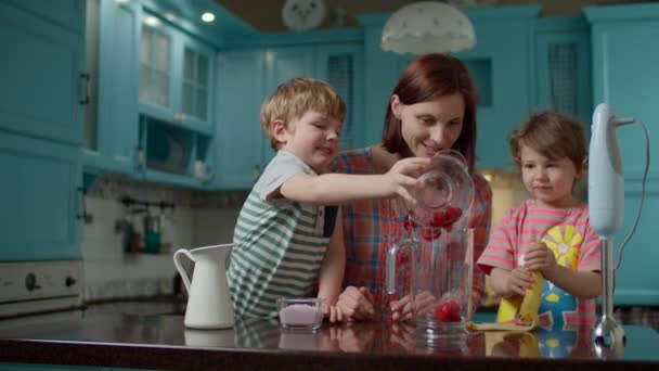 Feliz familia de madre y dos hijos cocinando batido de fresas, plátano, leche vegana y yogur con licuadora en casa cocina azul. Chico vertiendo bayas y yogur en un tazón, chica pelando plátano . — Vídeos de Stock