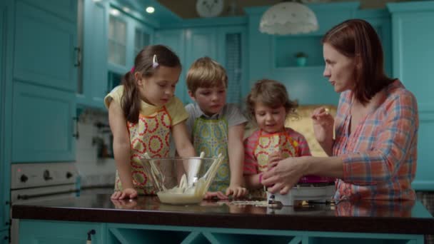 Eine junge Mutter mit drei Kindern in Schürzen backt zu Hause in der blauen Küche Waffeln. Kinder füllen Teig mit Löffeln in Waffelmaschine. Gemeinsam kochen. — Stockvideo