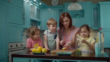 Üç çocuklu bir aile ev yapımı limonata, nane ve soda yapıyor. Genç anne, mavi mutfakta çocuklarla yemek pişiriyor.