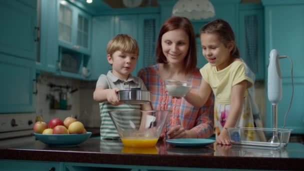 Feliz familia de madre y dos hijos cocinando tarta de manzana junto con huevos, azúcar, harina y manzanas en casa. Niño y niñas vertiendo harina para la masa en un tazón de vidrio . — Vídeos de Stock