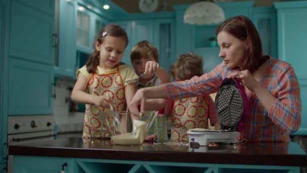 Eine junge Mutter mit drei Kindern in Schürzen backt zu Hause in der blauen Küche Waffeln. Kinder füllen Teig mit Löffeln in Waffelmaschine. Gemeinsam kochen. — Stockvideo