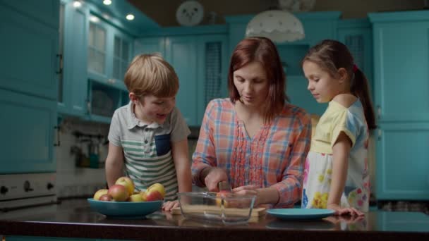 Feliz familia de madre y dos hijos cocinando tarta de manzana junto con huevos, azúcar, harina y manzanas en casa. Niño ayudando a mamá a cortar manzanas y poner en un tazón en la cocina azul . — Vídeos de Stock