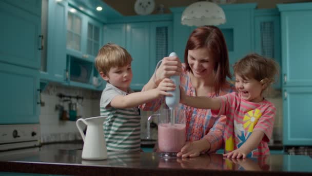 Glückliche Familie aus Mutter und zwei Kindern kochen Smoothie mit Erdbeeren, Banane, veganer Milch und Joghurt mit Mixer zu Hause blaue Küche. Junge und Mädchen helfen Mutter, einen gesunden Cocktail zu mixen. — Stockvideo