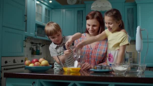 Feliz familia de madre y dos hijos cocinando tarta de manzana junto con huevos, azúcar, harina y manzanas en casa. Niño y niñas vertiendo azúcar para la masa en un tazón de vidrio . — Vídeo de stock