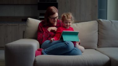 30 'lu yaşlarda, gözlüklü bir anne ve küçük bir kız evdeki kanepede oturup eğitim için tablet bilgisayar kullanıyor. Çevrimiçi uygulama kullanan aile. 