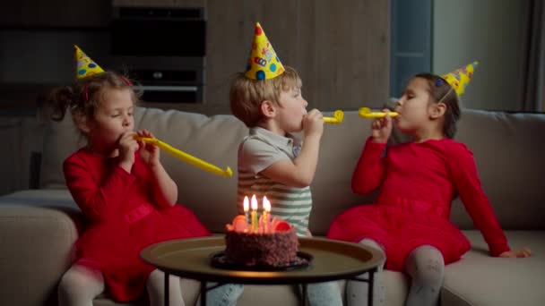 Trois enfants heureux dans des chapeaux d'anniversaire soufflant des pipes d'anniversaire les uns aux autres à la maison. Frères et sœurs célèbrent leur anniversaire avec un gâteau au chocolat et une bougie assis sur le canapé dans le salon . — Video