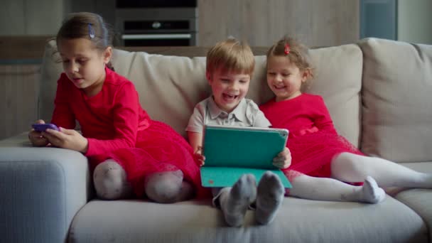 Niño y niña en edad preescolar usando tableta con hermana usando teléfono móvil sentado en el sofá en casa. Niños usando aplicaciones móviles en gadgets juntos . — Vídeo de stock