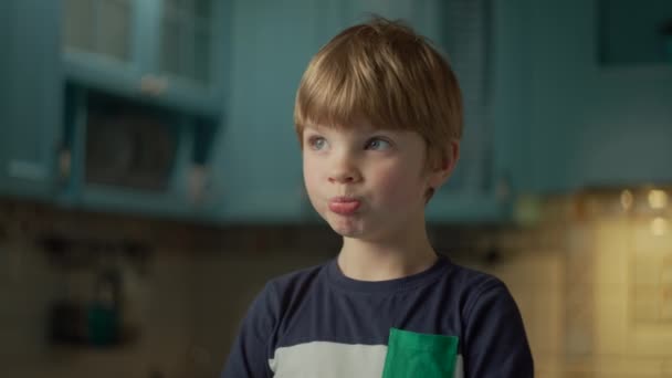 Porträt eines blonden Vorschulkindes, das gerne Schokoriegel isst, der auf einer blauen Küche steht. Kind beißt Schokolade in Händen ab. Kind isst gern Süßigkeiten. — Stockvideo