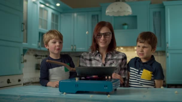 Щаслива сім'я молодої матері і двох синів готують поп-кукурудзу на сковороді на синій кухні вдома. Вибухи кукурудзи на гарячій сковороді при повільному русі . — стокове відео