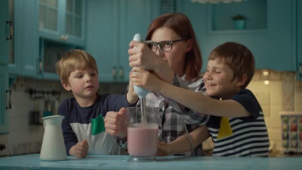 Gelukkige familie van moeder en twee kinderen koken smoothie van aardbeien, banaan, veganistische melk en yoghurt met blender thuis blauwe keuken. Familie geniet van blender samen en lachen. — Stockvideo