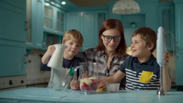Feliz familia de madre y dos hijos cocinando batido de fresas, plátano, leche vegana y yogur con licuadora en casa cocina azul. Niños vertiendo leche y yogur en un tazón . — Vídeos de Stock