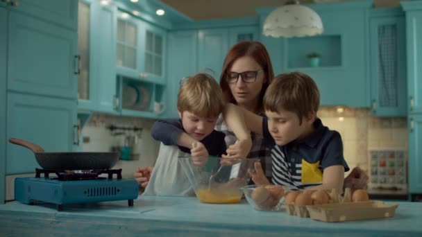 Boldog anya család és két fiú együtt főz rántottát a kék konyhán otthon. A gyerekek összekeverik a tojást villával az üvegtálban. Sütőserpenyő hordozható gáztűzhelyen. — Stock videók