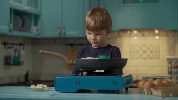 幼い就学前の少年は自宅の青いキッチンに一人で立って、スクランブルエッグを調理しました。子供はポータブルガスストーブの火をオンにし、動作するかどうかを確認します. — ストック動画