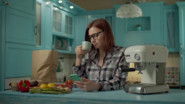 Mujer adulta joven en vasos de café y el uso de teléfono móvil de pie en la cocina azul en casa. Bolsa de papel y alimentos frescos de granja que ponen en la mesa de la cocina . — Vídeos de Stock