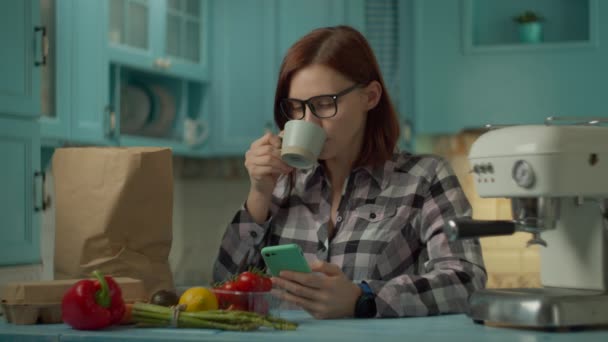 Jeune femme adulte dans des verres boire du café et en utilisant un téléphone portable debout sur la cuisine bleue à la maison. Sac en papier et nourriture fraîche à la ferme posé sur la table de cuisine. Gros plan — Video