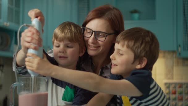 快乐的母亲和两个孩子在家里蓝色厨房用搅拌机煮草莓、香蕉、素食牛奶和酸奶。家人喜欢一起玩搅拌机，一起笑。靠近点 — 图库视频影像