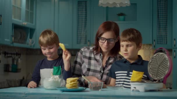 Jonge moeder uit de 30 die twee kinderen helpt om room en chocolade op zelfgemaakte wafels te doen die op een blauwe keuken staan. Kinderen eten graag zelfgemaakte snoepjes. Samen koken. — Stockvideo